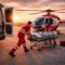 Emergency medical responders transfer a patient on a stretcher toward a medical evacuation helicopter at sunset, with an ambulance and medical equipment nearby.