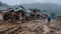 Kondisi banjir bandang di Desa Solimandungan II Kabupaten Bolaang Mongondow dengan rumah warga rusak, jalan dipenuhi lumpur, dan material kayu berserakan saat hujan masih turun.